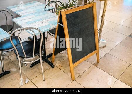 Lavagna vuota (spazio per menu) su pavimento piastrellato, tavoli e sedie vicino. Mockup per aggiungere il tuo testo. Foto Stock