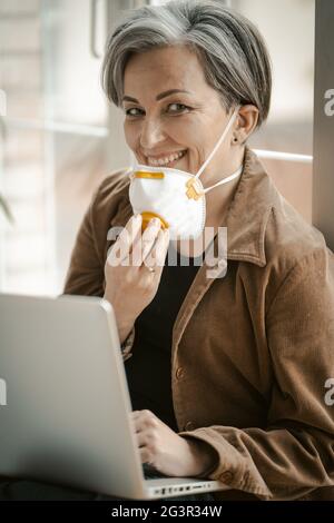 Donna dai capelli bianchi in età sorride togliendo la maschera e guardando la macchina fotografica. Donna sorridente con maschera protettiva funziona laptop Foto Stock