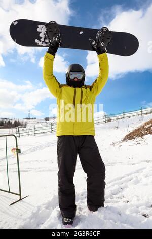 Happy snowboarder maschio caucasico indossando casco e occhiali, ritratto a lunghezza intera su cielo blu in montagna che sale su snowboard Foto Stock