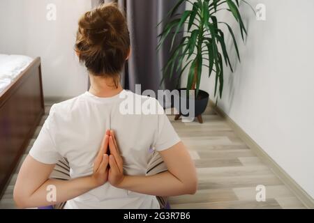 Giovane donna 20s che pratica yoga, facendo Namaste dietro, allenarsi, da solo a casa. Fit sana ragazza attiva sedersi sul tappetino sul pavimento in soggiorno, r Foto Stock