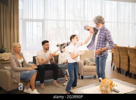 Nonno che ama ballare con la nipotina carina della bambina di fronte alla nonna e i genitori che si rilassano sul divano nel soggiorno a casa. Buona famiglia godendo del tempo libero e divertendosi insieme Foto Stock