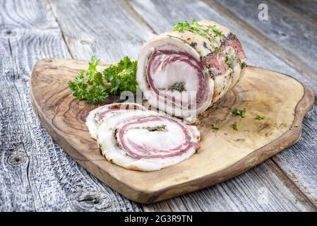 Tradizionale panchetta arrotolata italiana di carne di maiale affettata e come pezzo con erbe offerto come closeup su una rustica tavola di legno wi Foto Stock