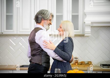 Coppia sposata senior in festa danzare in cucina elegante vicino a tavola con champagne a casa durante la festa di compleanno. Sorridendo marito guardando la moglie ed esprimendo il suo amore. Concetto di famiglia Foto Stock