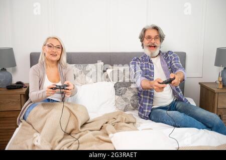 Divertirsi. Moglie e marito di coppia senior positivi che giocano a videogiochi con i joystick che competono e si godono il tempo libero insieme a casa, mentre comodamente seduti e rilassarsi su un letto spazioso Foto Stock