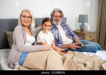 Ritratto di adorabile nipote di bambina e nonni senior che sorridono in macchina fotografica mentre si giocano i videogiochi seduti a letto e godendo del tempo libero di famiglia nel fine settimana a casa Foto Stock