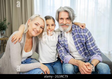 Ritratto di bella nipote bambina e sua pensionato sorridente bei nonni seduti sul divano in soggiorno a casa godendo di trascorrere il tempo insieme e condividere momenti speciali della famiglia Foto Stock