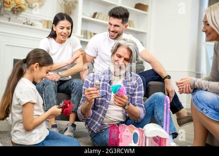 Piccola ragazza felice godendo del tempo libero con la famiglia, giocando con la madre del padre e i nonni mentre distendendosi insieme in soggiorno. Bella nipote che mostra la sua nuova casa delle bambole al nonno Foto Stock