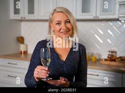 Ritratto di nonna sorridente in bel vestito festivo in piedi in elegante cucina che tiene un bicchiere di champagne che progetta di fare un brindisi e Wish Buon compleanno. Concetto di celebrazione Foto Stock