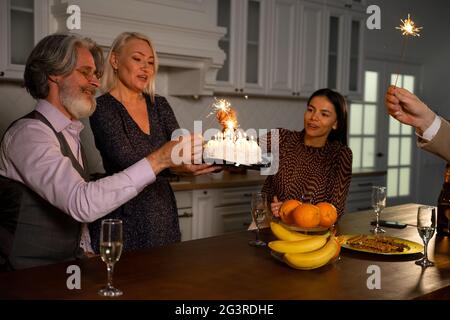 Buona famiglia festeggiando il compleanno di nonno eccitato, nonna portando torta con candele mentre gli ospiti tengono gli sparklers e gustando champagne in cucina a casa. Concetto di celebrazione Foto Stock