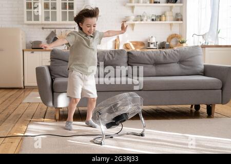 Il piccolo ragazzo gioca con forte vento soffiato dal ventilatore o dal ventilatore retrò a casa in un accogliente salotto da solo Foto Stock