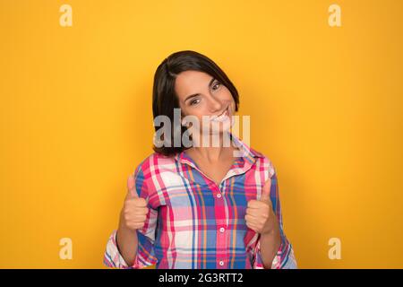 Allegra donna caucasica che mostra pollici in su. Giovane brunetta in taglio casual su sfondo giallo. Spazio di copia Foto Stock