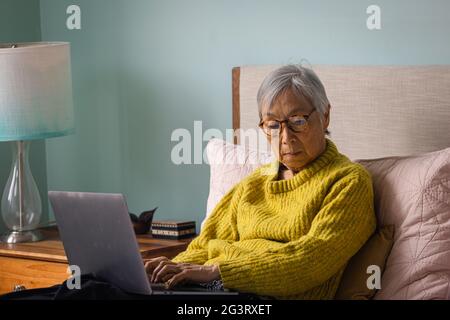 Donna anziana che usa il computer portatile mentre si siede in camera da letto a casa Foto Stock