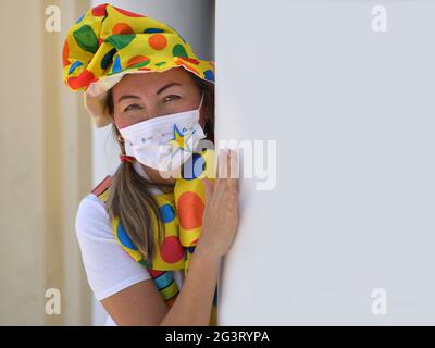 Il pagliaccio femminile con gli occhi sorridenti indossa un abito clown e una maschera chirurgica decorata, sbircia dietro un angolo durante la pandemia globale del coronavirus. Foto Stock