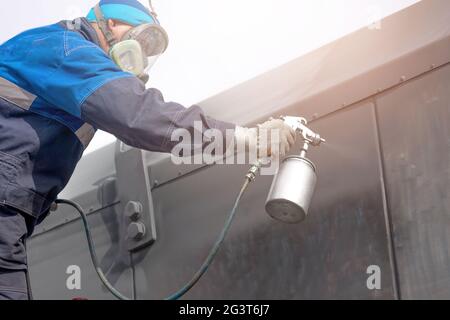 Lavoro industriale. Adescamento di prodotti metallici dalla pistola di compressione. Un operatore in tute e una maschera protettiva dipinge il corpo o Foto Stock