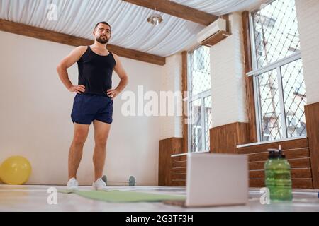 L'atleta si allena in palestra vuota o a casa davanti al computer portatile. Il giovane uomo isolato e motivato si scalda facendo spe Foto Stock