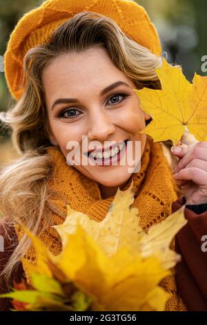 Primo piano. Giovane donna in berretto lavorato beige con foglie autunnali in mano e autunno giallo giardino o parco. Bella sorridente giovane Foto Stock