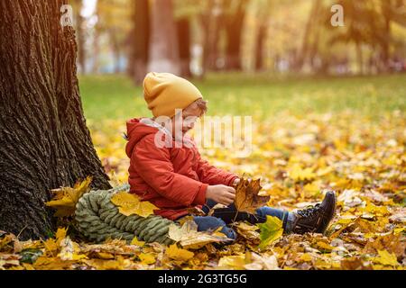 Piccolo ragazzo carino divertirsi all'aperto nel parco nel tempo di autunno seduto nelle foglie sotto un albero in una giacca calda autunno e una c Foto Stock