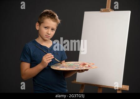 Cavaliere del pennello il ragazzo di 10 anni guarda la macchina fotografica che tiene una tavolozza con le pitture sullo sfondo di un cavalletto con tela. SCH Foto Stock