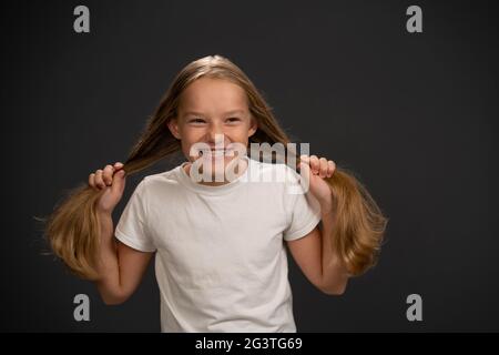 Felice sorridente e tenendo due code pony 8,10 anni ragazza tenendo i suoi capelli indossando t shirt bianca sorridendo un po 'grinning a. Foto Stock