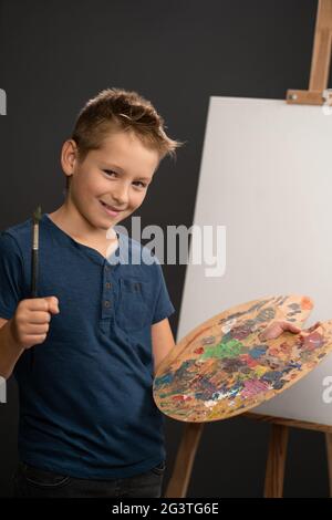 Affascinante caucasico ragazzo di 10 anni guarda la macchina fotografica che tiene una tavolozza con le pitture sullo sfondo di un cavalletto con tela. Sho Foto Stock