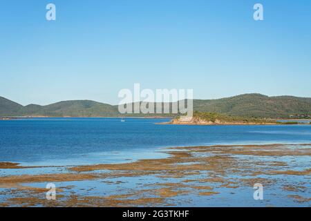 Vista dalla cima di una collina sull'oceano blu a bassa marea con uno yacht ormeggiato al largo sotto un cielo blu chiaro e uno sfondo di montagna Foto Stock