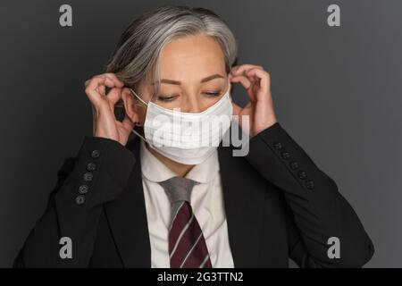 Donna d'affari di mezza età con capelli grigi che si appone su una maschera medica protettiva bianca che guarda verso il basso. Ritratto della donna anziana moderna Foto Stock