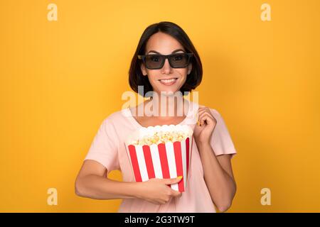 Secchio di popcorn e occhiali stereo 3D nelle mani di una giovane donna di brunette. Concetto di cinema. Isolato su sfondo giallo Foto Stock