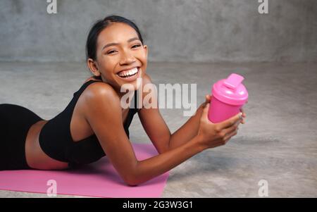 Felice donna sorridente sana che tiene le vibrazioni della proteina che si rilassa sul tappeto di yoga rosa in palestra Foto Stock