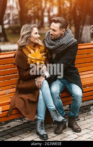 Vista frontale di una coppia innamorata seduta sulla panchina abbracciata nel parco con stemmi e sciarpe che raccolgono un bouquet di congedo caduto Foto Stock