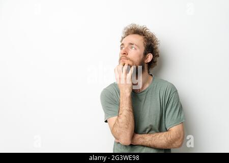 Barba grattata e appicante giovane uomo con le mani piegate. Bell'uomo giovane con capelli ricci in t-shirt oliva guardando lontano isolat Foto Stock