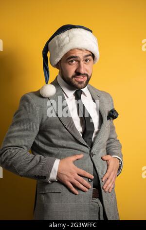 L'eccesso di cibo, l'alimentazione impropria durante le feste di Capodanno bell'uomo sopportato in cappello blu di Santa e vestito grigio, guardando la camma Foto Stock