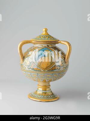 Vaso con tre orecchie e un coperchio, dipinto multicolore con spittsavals all'interno del quale l'uomo ritratti, e fiore e foglia raffinano. Vaso sferico di maiolica dipinta multicolore, ad un piede alto. Il vaso ha tre orecchie e un coperchio. Ci sono tre obal attaccante sul ventre in cui un ritratto di un uomo spostato con capelli lunghi. Fiore e foglie sono dipinti tra gli ovali. Il vaso è verniciato in lucentezza gialla e in blu (circonferenza) su bianco. Foto Stock