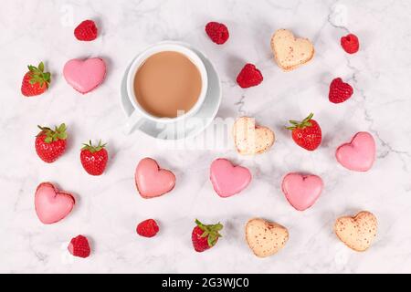 I dolci francesi a forma di cuore rosa e beige accanto alla tazza da caffè e alla frutta alla fragola Foto Stock