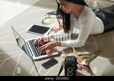 Contenuto giovane fotografa asiatica giacente sul tappeto tra fiori, dispositivi e foto fotografiche e ritocchi sul laptop Foto Stock