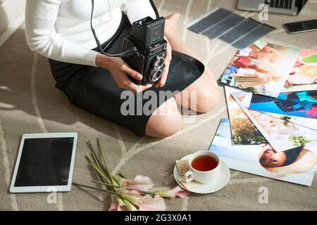Primo piano di una fotografa femminile irriconoscibile in gonna nera seduta in ginocchia con una fotocamera vintage tra foto colorate luminose sul tappeto Foto Stock
