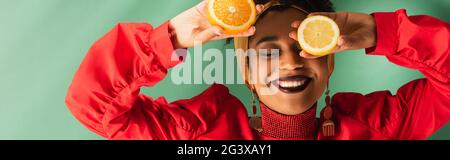 sorridente giovane afroamericana tenendo mezzo taglio di limone e arancio vicino faccia su verde, banner Foto Stock