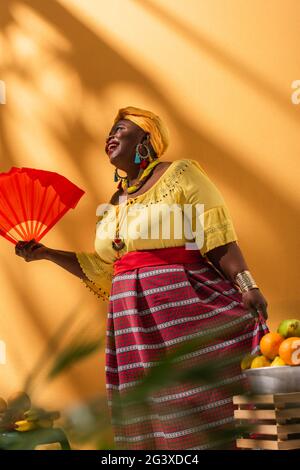 vista ad angolo basso di una donna afro-americana di media età positiva che vende frutta e tiene la ventola sull'arancione Foto Stock