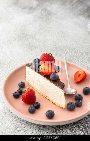 Un pezzo di cheesecake coperto di cioccolato con mirtilli e fragole primo piano su un piatto rosa con un cucchiaio da dessert Foto Stock