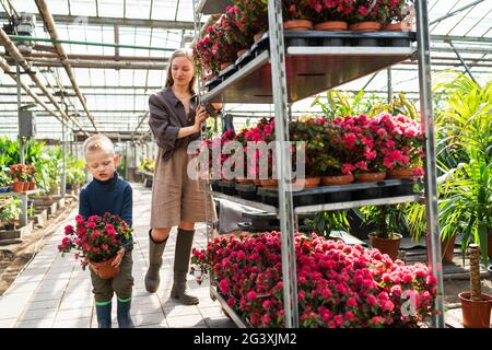 Il ragazzo scontento porta un vaso di fiori e la madre tira un carro con piante da dietro Foto Stock