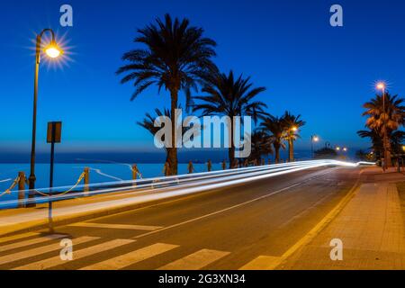 Una lunga esposizione vista notturna della strada fronte oceano con linee chiare da auto e palme di passaggio in silhouette e oceano in Foto Stock