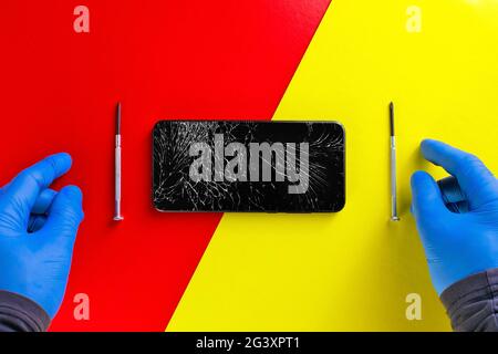Le mani guastate di un riparatore di hardware accanto a cacciaviti e un telefono rotto su uno sfondo giallo-rosso. Vista dall'alto Foto Stock