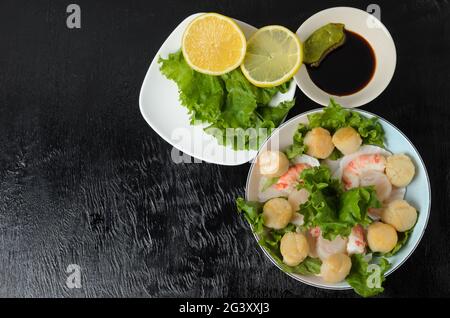 Carne di capesante fritta con gamberi bolliti in un piatto di ceramica con lattuga, limone, salsa di soia e wasabi su fondo scuro. Messa a fuoco selettiva. Foto Stock