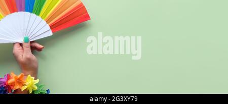 Banner di orgoglio della comunità LGBTQ. Mano con bandiera arcobaleno, ghirlanda di fiori e unghie finte in colori di arcobaleno. Foto concettuale. Piatto su blu menta Foto Stock