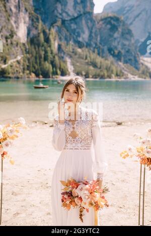 Bella sposa in un abito bianco con maniche e pizzo, con un bouquet giallo autunno sullo sfondo dell'arco per la cerimonia, a. Foto Stock