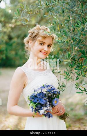 Una sposa tenera con un bouquet di fiori blu in le sue mani sono in piedi da rami di olive verdi in un boschetto e sorrisi Foto Stock