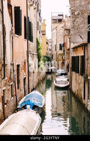 Barche ormeggiate alle pareti delle case in acqua a Venezia. Stretto canale desertato tra le case, acque marine azzurre ai piedi di Foto Stock