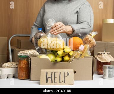 Donna in un maglione grigio mette in una scatola di cartone vari alimenti, frutta, pasta, olio di girasole in una bottiglia di plastica Foto Stock