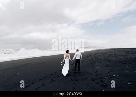 Una coppia di nozze sta camminando lungo la spiaggia nera di Vic. Spiaggia sabbiosa con sabbia nera sulle rive dell'Oceano Atlantico. Sposa Foto Stock
