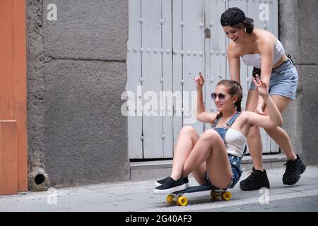 Una donna che spinge la sua amica da dietro su uno skateboard. Due amiche che si divertono in strada. Foto Stock