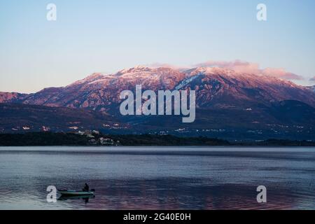 Un pescatore sta pescando in una barca nella baia di Cattaro, sullo sfondo del monte Lovcen coperto di neve al tramonto. Foto Stock